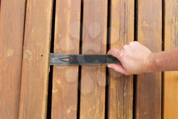 Horizontal photo of hand with pry bar lifting up old cedar wood board on outdoor wooden deck 
