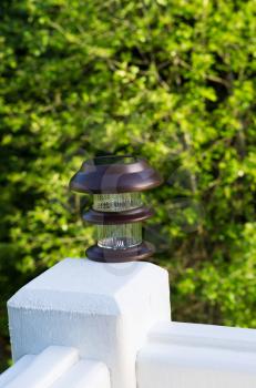Vertical photo of solar lamp, during day light, on deck post with green trees in background 