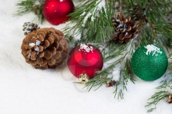 Horizontal view of Christmas ornaments, resting in white snow, with evergreen tree branch in background 