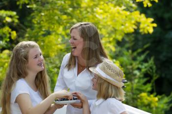 Side view of mother laughing with her daughters while eating fresh blue berries outdoors on patio with woods in background 

