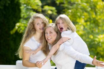 Front view, looking forward, of mother holding her younger daughter with older daughter hugging them while outdoors on patio with blurred out woods in background 