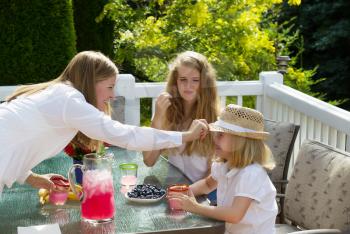Side view of happy mother putting hat on younger daughter while eating breakfast outdoors, during summer time, on patio with woods in background 