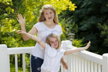 Front view, looking forward, of two sisters waving their arms while outdoors on patio with blurred out trees in background 