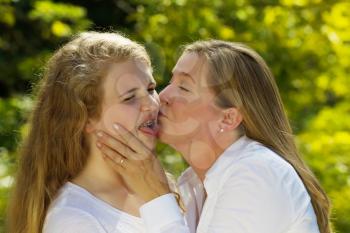Side view of mother kissing her teenage daughter, expressing herself in a negative way, while holding her face in her hands during outing outdoors on patio with blurred out woods in background 