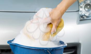 Closeup of female hand holding soapy sponge out of the bucket with car in background 