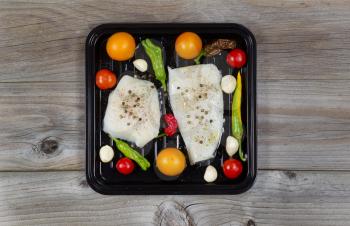 Top view of fresh raw Cod Fish Fillets in cooking pan with tomatoes, garlic, peppers and peppercorn salt on rustic wooden boards 