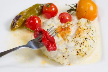 Closeup of fork and red pepper with baked stuffed sole fish, tomatoes, green pepper in sauce on white plate 