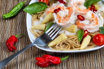 Closeup front view of stainless steel fork holding single shrimp with Alfredo Pasta dinner placed on white plate and natural bamboo mat underneath 