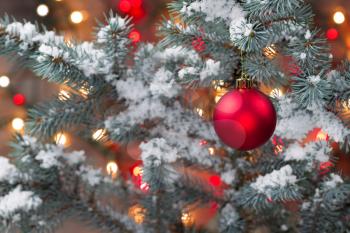 Closeup horizontal image of a single red Christmas ornament hanging from snow covered pine tree with glowing lights in background 