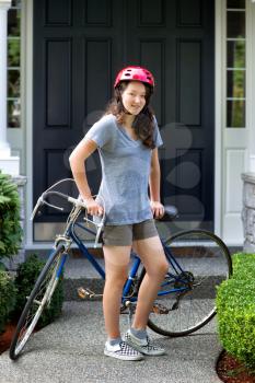 Vertical image of teenage girl, looking forward, while resting against her bicycle with home in background 