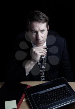 Vertical image of business man, looking at computer screen while holding pen, working late with black background 
