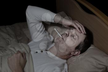Horizontal image of mature man, with high fever, lying down in bed testing his temperature 