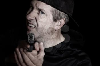 Closeup front view of mature man, looking forward and wearing baseball cap backwards, with gun in hand on dark background 