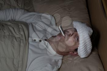 Horizontal image of mature man, with wet towel on forehead, lying down in bed testing his temperature 