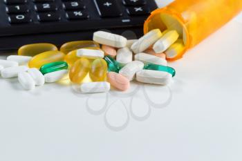 Close up of various medicine pills and tablets spilling out of plastic bottle in front of partial calculator on white background 