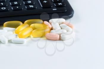 Close up of various medicine pills and tablets in front of partial calculator on white background 