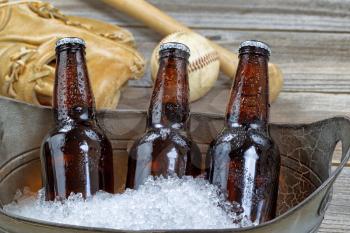Close up of three brown cold bottled beers, crushed ice in metal container, and baseball equipment in background on rustic wood