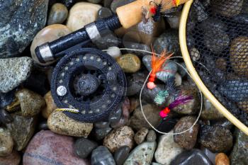 Overhead view of fishing fly reel, landing net and assorted flies on wet river bed stones