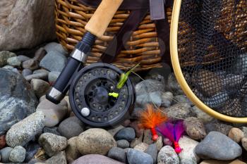 Close up of  fishing fly reel, landing net, creel and assorted flies on dry river bed rocks 