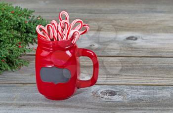 Close up of a red mug filled with holiday candy canes with partial Christmas tree on rustic wood 