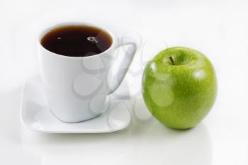 Close up of a single pristine green apple with a cup of fresh black coffee on a soft white table with light reflections. Focus on front top of objects.  