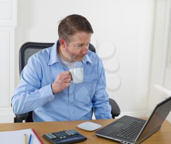 Mature man holding cup of coffee while looking at laptop screen, calculator, pencils, and notepad on desk. Background is white walls.  