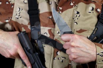 Close up horizontal image of hand holding knife with armed male soldier in background. Focus on knife and hand. 
