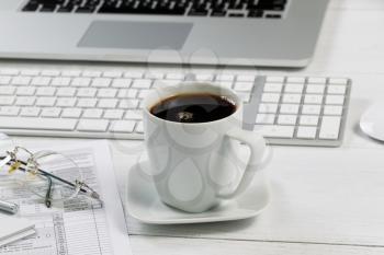 Close up of a fresh cup of black coffee with laptop, keyboard, pen, mouse, reading glasses and tax forms in background on white desk. Focus on front lip of coffee cup. 