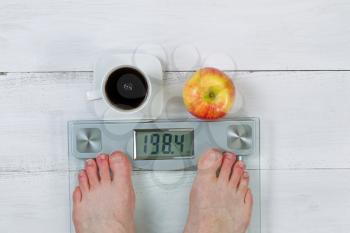 Weight scale, displaying body weight, with bare male feet and whole apple with black coffee in front  on white wooden floor 
