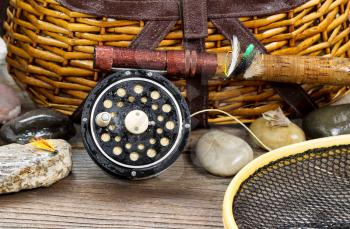 Close up of a wet antique fly fishing reel, rod, landing net, artificial flies and rocks in front of creel with rustic wood underneath. Layout in horizontal format.