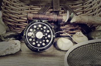 Vintage concept with grain of a wet antique fly fishing reel, rod, landing net, artificial flies and rocks in front of creel with rustic wood underneath. Layout in horizontal format.