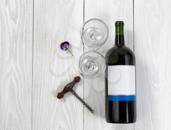 Overhead angled view of a large bottle of red wine, drinking glasses, flower and antique corkscrew on white wooden boards. 