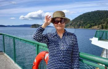 Senior woman, looking forward, wearing hat and sun glasses while standing on boat dock with ocean and blue sky in background 