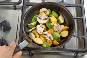 Close up of hand holding frying pan, focus on food, while cooking vegetables in pan on top of stove.