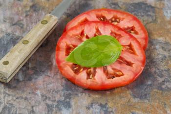 Close up view of single large basil leaf, focus on leaf, resting on top of a freshly sliced tomato with knife. Real stone cutting board. 