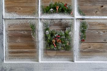 Snow covered window with decorative Christmas wreath on rustic wooden boards in background. Focus on window glass and sills. 
