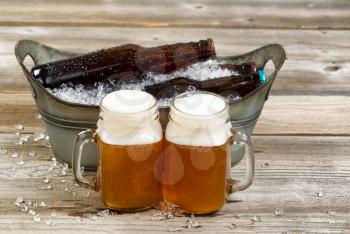 Two pints of frosty cold beer with metal bucket and ice in background on rustic wooden boards. 