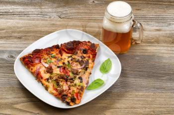 Two slices of freshly baked pizza in a white plate with a pint of justly poured beer setting on a rustic wooden table.