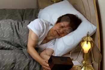 Senior woman looking at alarm clock during night time. Insomnia concept. 
