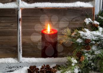 Decorated window with glowing red candle, selective focus on flame and top part of candle, pine tree, cones and snow outside. Christmas concept.