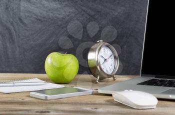 School desktop with laptop, mouse, clock, cell phone, notebook, pen and green apple in front of blackboard. Layout in horizontal format with copy space. 