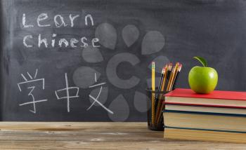 Front view of rustic desk for student learning Chinese with books, pencils and a green apple in front of chalkboard with Mandarin text. 