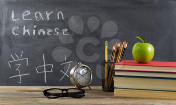 Student learning Chinese with books, pencils, clock, reading glasses and an apple in front of chalkboard with Mandarin text. 