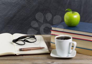 Close up of a rustic desktop of teacher with books, paper, pen, coffee, reading glasses a long with a green apple in front of chalkboard. Layout in horizontal format with copy space. 