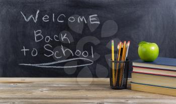 Old wooden desktop with basic school supplies and welcome back to school text on blackboard for students. Layout in horizontal format.