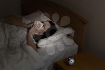 Woman wiping nose with tissue while trying to sleep. Select light and focus on woman and clock with darker background. Sickness at night time concept. 