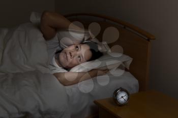 Woman with insomnia covering her ears with pillow while eyes open. Select light and focus on woman with darker background for night time concept.