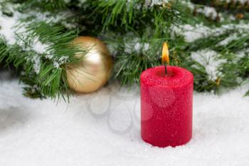 Close up of burning red candle with evergreen branches and gold ornaments covered in snow. Selective focus on front upper part of candle and flame with Christmas concept. 