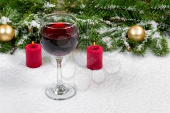 Close up of a glass of red wine with burning red candles, evergreen branches and gold ornaments covered in snow. Selective focus on front upper part of wine glass with holiday concept. 
