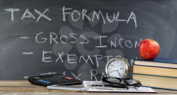 Desktop with tax form, books, apple, reading glasses, clock, pens, chalkboard, and calculator. Focus on front part of desk objects with classroom concept. 